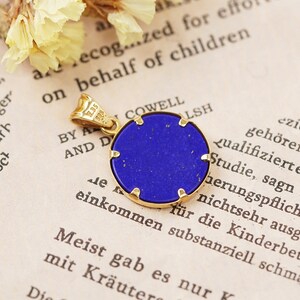 May include: A round pendant with a deep blue stone set in a gold-colored frame. The pendant has a small loop for a chain and is displayed on a vintage book page with text and dried flowers.