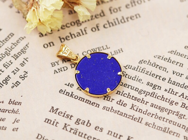May include: A round pendant with a deep blue stone set in a gold-colored frame. The pendant has a small loop for a chain and is displayed on a vintage book page with text and dried flowers.