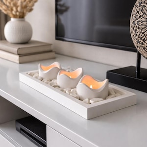 Op de afbeelding: Een wit rechthoekig dienblad met drie witte kaarshouders, elk met een brandende kaars. De lade is gevuld met kleine witte stenen. De kaarsen staan op een wit oppervlak, met een vaas en boeken op de achtergrond.