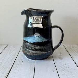 May include: A black ceramic pitcher with a handle and spout. The pitcher features a landscape design with blue and brown tones. A white label is affixed to the front of the pitcher.