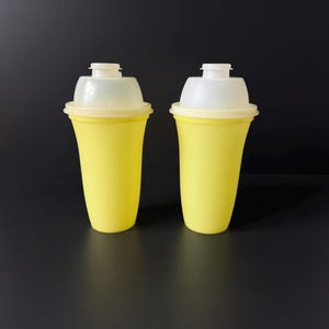 May include: Two matching yellow plastic shaker cups with translucent white lids. Each cup features a tapered cylindrical body and a secure, snap-on white cap at the top. The containers are identical in design, color, and size, standing upright.
