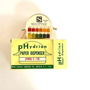May include: A pHydrion paper dispenser box with yellow packaging and green text, accompanied by a clear plastic test kit featuring a color-coded pH scale from 1 to 12, labeled Shaklee Corporation, Emeryville, CA 94608 and Micro Essential Laboratory Inc.