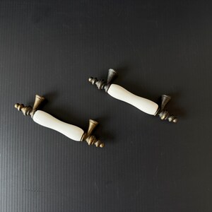 May include: Two drawer pulls featuring a smooth, white, cylindrical center section made of ceramic material, flanked by ornate, dark, antique-style brass metal ends with a tapered, conical shape and decorative ringed detailing at the base.