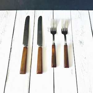 May include: Set of vintage silverware including two knives and two forks. The knives have dark blades and brown handles. The forks have silver tines and brown handles. The silverware is arranged on a white wooden surface.