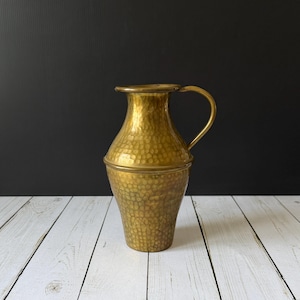 May include: A hammered brass pitcher with a handle. The pitcher has a wide base that tapers to a narrower neck and a flared rim. The surface of the pitcher is covered in a textured pattern. The pitcher is a warm gold color.