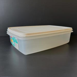 May include: A rectangular, translucent plastic food storage container with a white lid. The container is empty and sits on a dark surface. A label is visible on the side of the container.