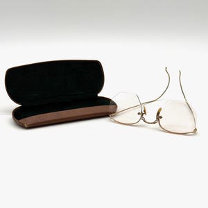 May include: A pair of vintage rimless eyeglasses with a gold-colored metal frame and clear lenses. The glasses are next to an open brown leather eyeglasses case with a black velvet interior. The case is open.