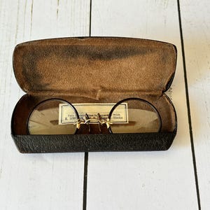 May include: Vintage round eyeglasses with a gold-tone bridge and temples, resting inside an open, worn leather case. The lenses are clear, and a small paper label is visible. The case is dark brown with a textured interior.