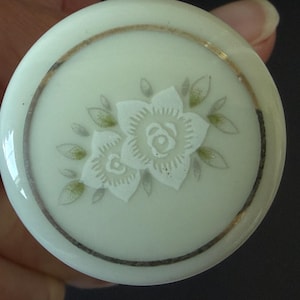 May include: A round, off-white ceramic knob featuring a central embossed floral design with two white flowers and small green leaves. A thin, metallic gold-colored circular border encircles the floral motif, set against a smooth, glossy finish.