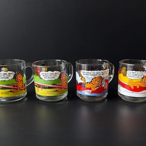 May include: Four clear glass mugs with handles, each featuring a Garfield cartoon character design. The mugs have different quotes and colorful backgrounds, including yellow, red, and blue. The mugs are suitable for beverages.