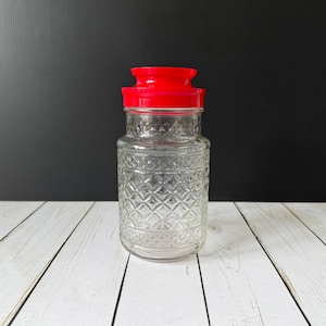 May include: A clear glass jar with a red lid. The jar has a diamond pattern etched into the glass. The jar is sitting on a white wooden surface with a black background.