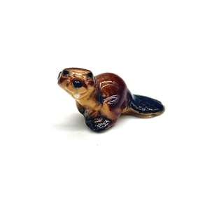 May include: A small ceramic beaver figurine. The beaver has a brown body, a dark blue tail, and black eyes. The figurine is on a white background.