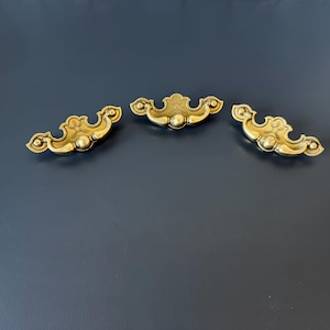May include: Three matching gold-toned metal drawer pulls featuring a curved, ornate design with a central circular knob and decorative scrollwork on the sides. Each handle has a symmetrical shape with pointed ends and a polished metallic finish.
