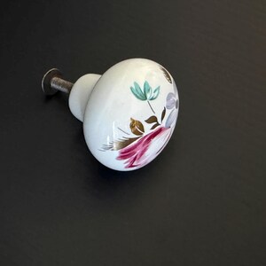 May include: A white ceramic cabinet knob with a rounded, smooth surface featuring a painted floral design in shades of pink, teal, and purple with brown accents. The knob includes a silver-toned metal screw attachment extending from the back.