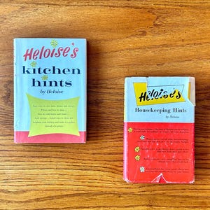 May include: Two vintage books titled "Heloise's Kitchen Hints" and "Heloise's Housekeeping Hints" are displayed on a wooden surface. The books have colorful covers with text in red, yellow, and blue. The kitchen hints book has a light blue background.