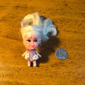 May include: A small doll with blonde hair and blue eyes, wearing a purple dress with white flower accents. A dime is next to the doll for size comparison. The doll is on a wooden surface.
