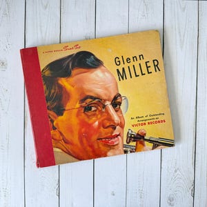 May include: A vintage Glenn Miller record album with a red spine and a yellow cover featuring a painted portrait of a man wearing glasses and holding a trumpet. The text reads Glenn Miller, A Victor Musical Smart Set, and An Album of Outstanding Arrangements on Victor Records.