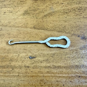 May include: A silver-colored latch hook tool with a curved hook on one end and a textured loop on the other. The tool is lying on a wooden surface.