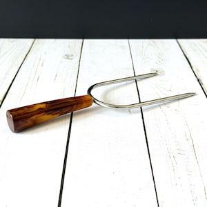 May include: A vintage carving fork with a brown and amber marbled handle and two stainless steel tines. The fork is designed for serving meats and other foods. The handle is made of a durable material and is comfortable to hold.