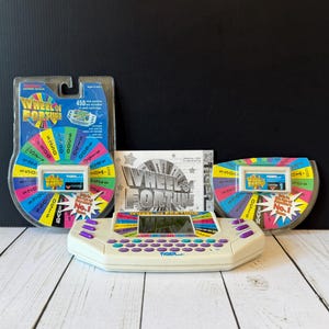 May include: A Tiger Electronics Wheel of Fortune handheld electronic game console with a QWERTY keyboard, purple buttons, and a screen. Included are two blister-packed game cartridges featuring a colorful prize wheel graphic and the text Wheel of Fortune.