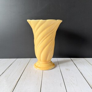 May include: A pale yellow ceramic vase featuring a distinct spiral or swirl pattern along its body. The vase has a wide, flared rim and a circular pedestal base, with a smooth, glossy finish that reflects light across its curved, textured surface.