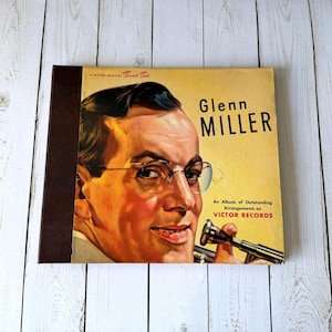 May include: Vintage Glenn Miller album cover featuring a portrait of the musician in glasses, holding a trumpet. The album cover is yellow with the title "Glenn Miller" and the text "An Album of Outstanding Arrangements on VICTOR RECORDS."