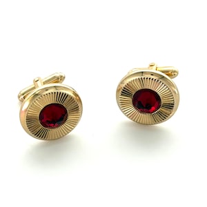 May include: A pair of gold-toned cufflinks with a circular design. Each cufflink features a vibrant red gemstone at the center, surrounded by a sunburst pattern. The cufflinks have a classic, elegant style.