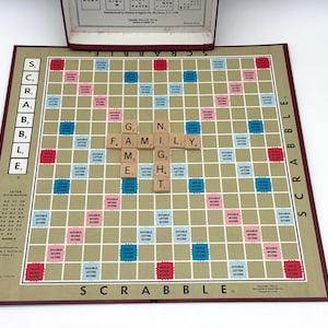 May include: A vintage Scrabble board game with the word "FAMILY" spelled out. The board is tan with red, blue, and pink squares. The game's box is open in the background. The word "SCRABBLE" is printed on the board.