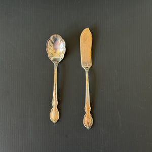 May include: A silver spoon with a scalloped bowl and a butter knife with a wooden handle are displayed on a dark background. The spoon and knife have ornate handles and are likely vintage or antique.