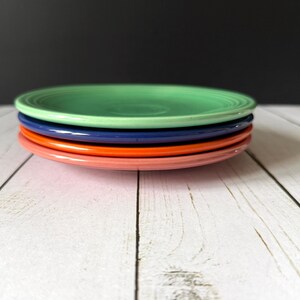 May include: A stack of four colorful ceramic plates. The top plate is green, followed by blue, orange, and pink. The plates are arranged on a white wooden surface with a dark background. These plates are suitable for serving food.