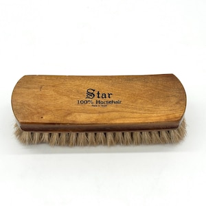 May include: A wooden brush with a curved handle and natural horsehair bristles. The brush is light brown with the text "Star 100% Horsehair Made in Brazil" printed on the handle. The bristles are a light tan color.