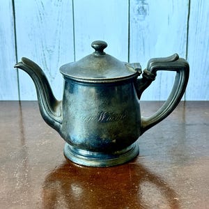 May include: Antique silver-toned teapot with a lid and handle. The teapot has a spout and a small base. The word "Wayne Winkle" is engraved on the side. The teapot is sitting on a brown surface.