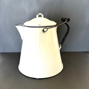 May include: Cream-colored enamelware coffee pot with a lid and black trim. The pot has a black handle and a spout for pouring. The vintage kitchenware is likely used for brewing coffee or tea.