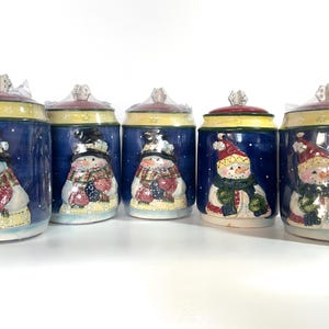 May include: Set of five ceramic canisters with snowman designs. Each canister features a different snowman with a blue background and white accents. The canisters are perfect for storing sugar, flour, coffee, tea, or other kitchen staples.