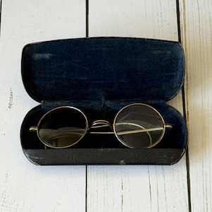 May include: Open black eyeglass case with a pair of round, gold-rimmed eyeglasses. The lenses are dark, and the case has a dark blue velvet interior. The case is open, revealing the glasses inside.