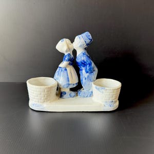 May include: A ceramic figurine depicting a kissing couple in traditional Dutch attire, with blue and white details. The figurine has two small, basket-like containers on either side of the couple, and is set on a white base.