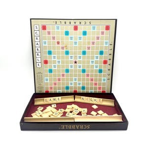 May include: A Scrabble board game set, open to display the game board, letter tiles, and tile racks. The board is tan with colored squares. The game's name is printed on the board and box.