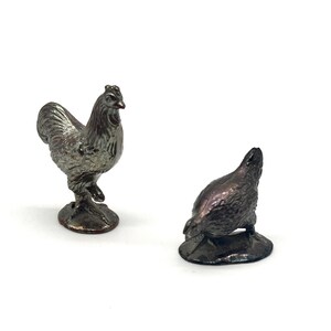 May include: Two small, metallic rooster and hen figurines. The rooster stands upright, while the hen is in a resting position. Both are detailed with textured surfaces and are set against a white background.