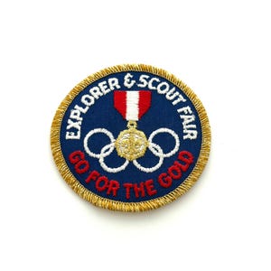 May include: A round, embroidered patch with the text "EXPLORER & SCOUT FAIR" and "GO FOR THE GOLD" in white and red lettering. The patch features a gold medal with Olympic rings and a red and white ribbon, all set against a navy blue background.