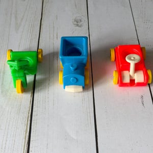 May include: Three colorful toy vehicles: a green scooter, a blue truck, and a red car. Each toy has yellow wheels and is made of plastic. The toys are arranged on a light-colored wooden surface.