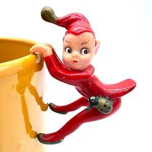 May include: A yellow ceramic mug featuring a three-dimensional red elf figure attached to the side. The elf has a pointed hat, large eyes, and a small ladybug accessory on its hip, appearing to climb the exterior wall of the bright yellow cup.