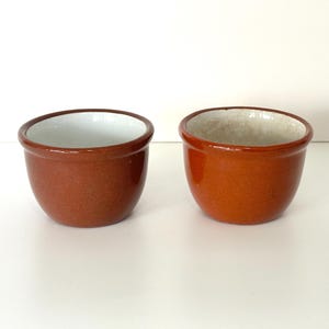 May include: Two small, terracotta-colored ceramic bowls with white glazed interiors. The bowls are round and have a slightly flared lip.