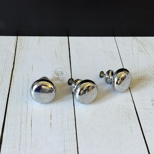 May include: Three round cabinet knobs with a polished chrome finish. Each knob features a smooth, reflective surface and a short metal stem with a screw attachment extending from the back, designed for mounting onto furniture or cabinetry doors.