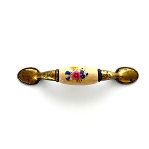 May include: A drawer pull featuring a central cream-colored ceramic barrel with a painted floral design in pink, blue, and yellow. The handle has weathered, tarnished brass-toned metal ends with a distressed finish on both sides of the ceramic.