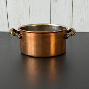 May include: A round copper cooking pot with a polished metallic exterior and two curved brass handles on opposite sides. The interior of the pot features a silver-toned lining, and the rim is smooth and circular, showcasing a classic design.