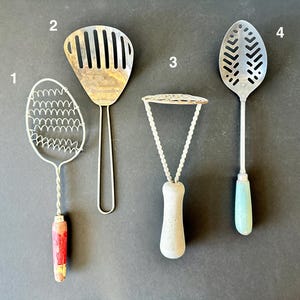 May include: Four vintage kitchen utensils are displayed against a dark background. They include a wire egg whisk with a painted handle, a slotted spatula, a potato masher, and a slotted spoon with a light blue handle. These tools are likely used for cooking.