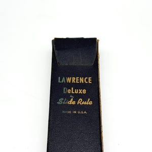 May include: A dark blue rectangular case with a flap closure. The front features gold-colored text that reads LAWRENCE, DeLuxe, Slide Rule, and MADE IN U.S.A. The case has a textured surface and visible stitching along the side edge.