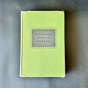 May include: A vintage green hardcover book titled "The Good Housekeeping Needlecraft Encyclopedia." The book has a light green cover with a gray and white label. The book is on a gray surface.
