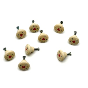 May include: A collection of nine round, cream-colored cabinet knobs, each featuring a central red heart design surrounded by small black floral accents. Each knob includes a metal screw extending from the back for attachment to furniture.