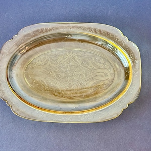 May include: An oval, clear glass serving tray with a gold-colored rim. The tray features an embossed floral design around the edges and a central diamond-shaped pattern. The tray is approximately 12 inches long and 8 inches wide.